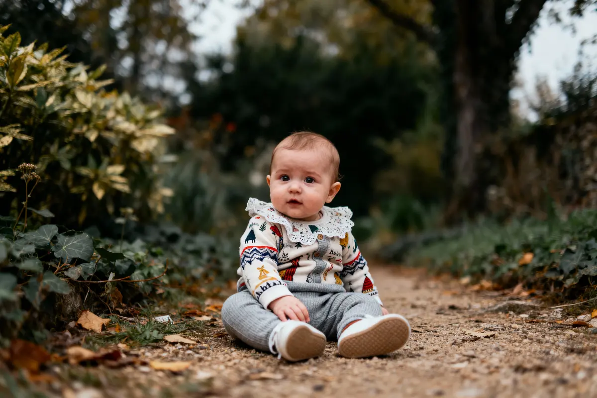Bébé assis sur un chemin forestier, portant un pull coloré avec dentelle, entouré de feuillage automnal.