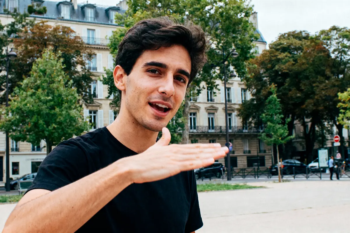 Un homme en t-shirt noir dans un parc urbain, gesticulant devant des arbres et immeubles.
