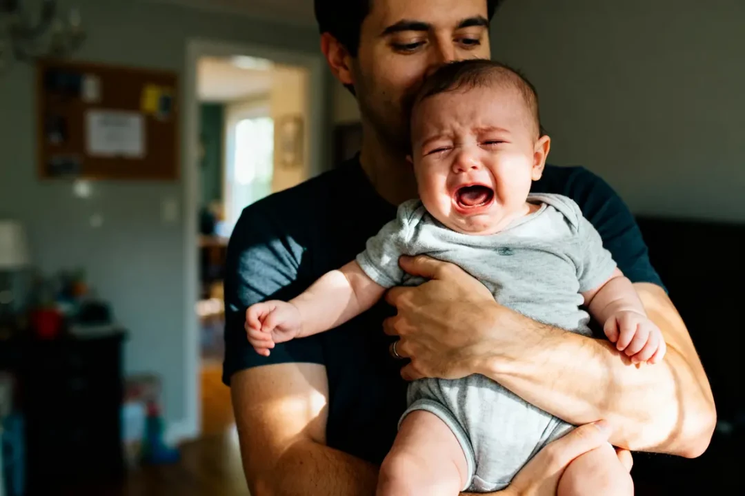 Un homme tient un bébé pleurant, vêtu d'un body gris, dans une pièce éclairée.