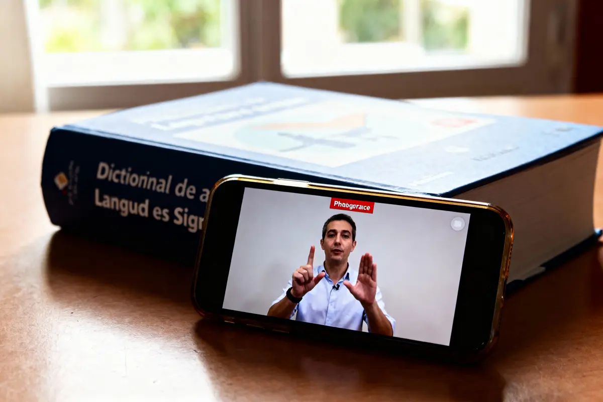 Homme communiquant en langue des signes sur smartphone, livre du dictionnaire de langue des signes en arrière-plan.