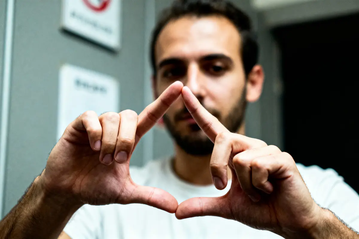 Homme montrant un triangle avec ses mains, en arrière-plan flou et panneaux muraux visibles.