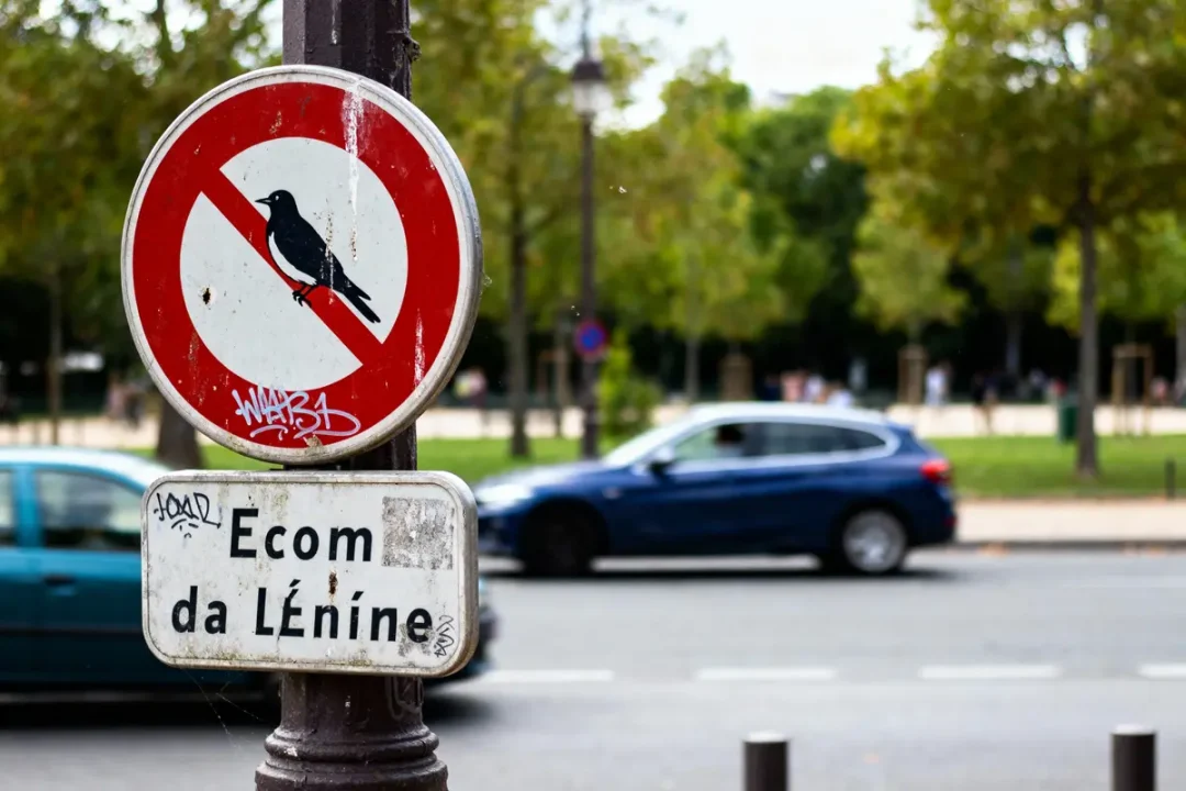 Panneau d'interdiction avec oiseau barré, graffiti, voitures et arbres en arrière-plan.