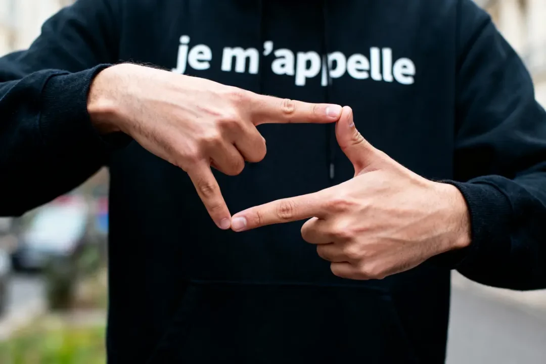 Personne en sweat noir, fait un cadre avec les doigts, texte "je m'appelle" visible sur le sweat.