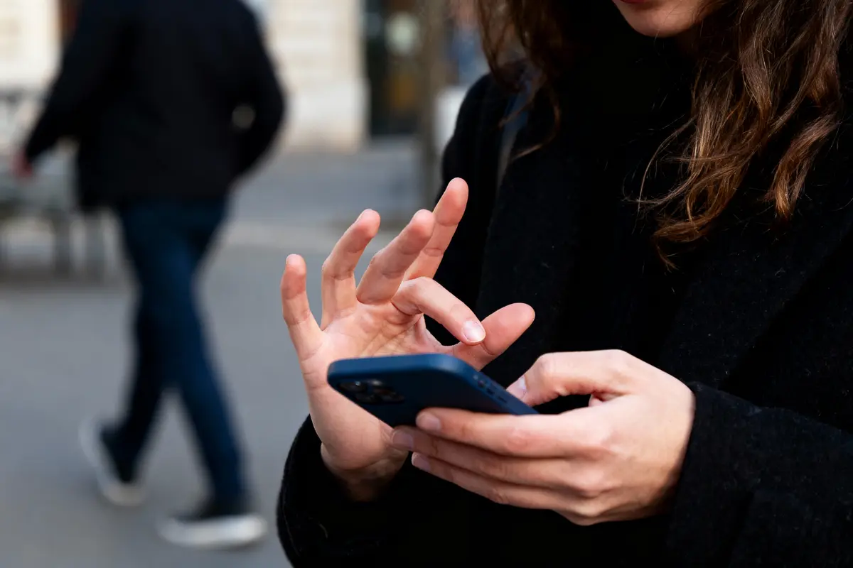 Personne en manteau noir utilisant un smartphone bleu en extérieur, avec un piéton flou en arrière-plan.
