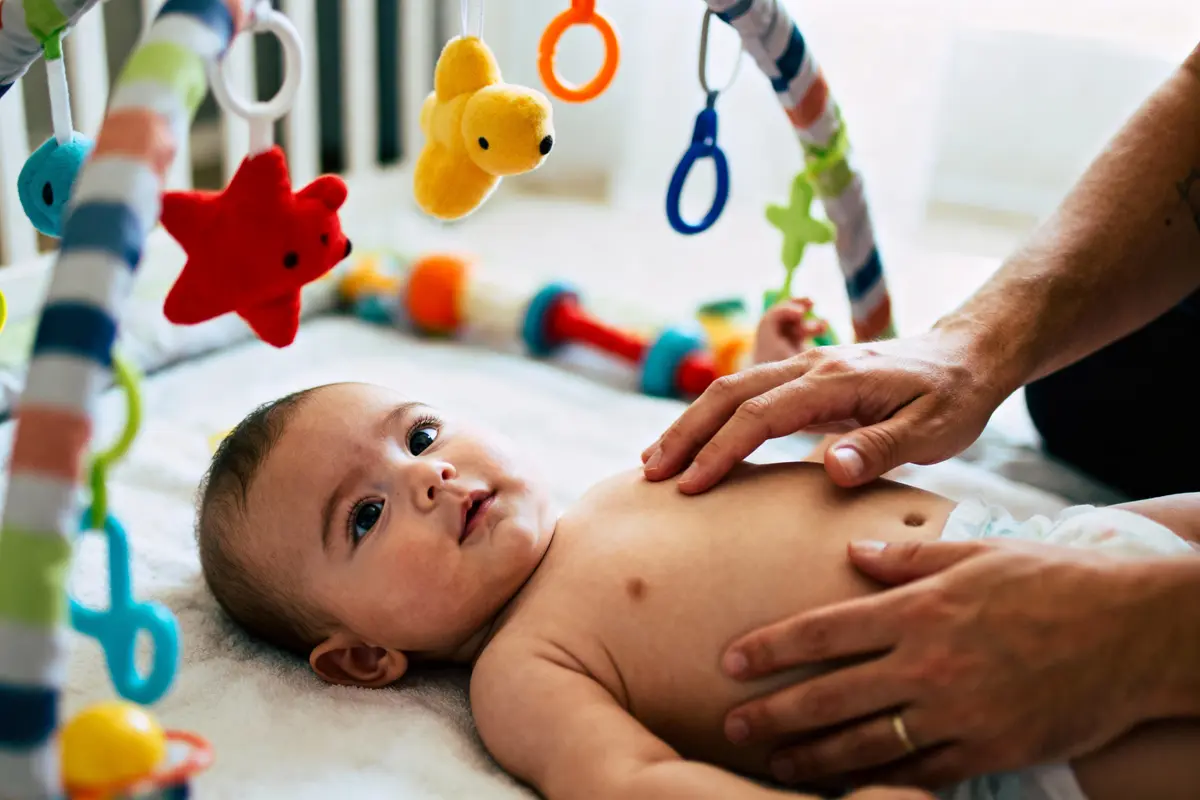 Bébé couché sous arche de jeu colorée, mains d'adulte touchant doucement son ventre.