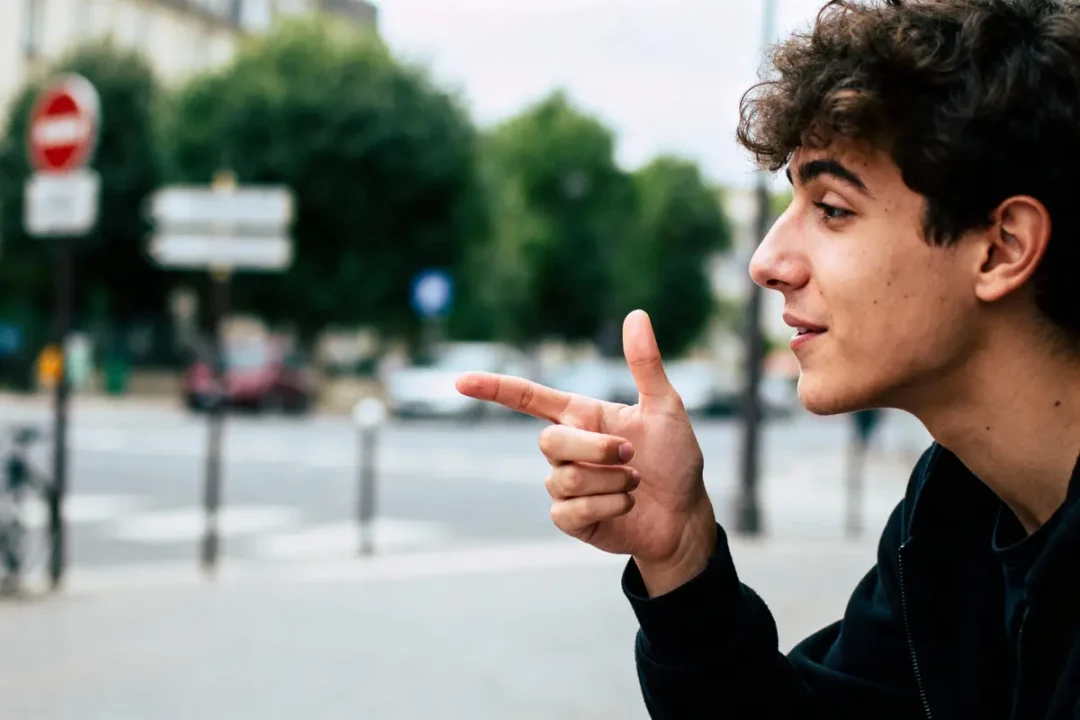 Jeune homme souriant, profil, faisant un geste de la main en extérieur urbain flou, arbres en arrière-plan.