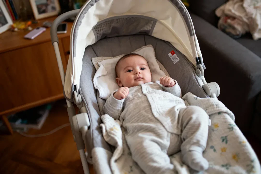 Bébé en pyjama gris allongé dans un landau, intérieur confortable avec meubles et objets divers autour.