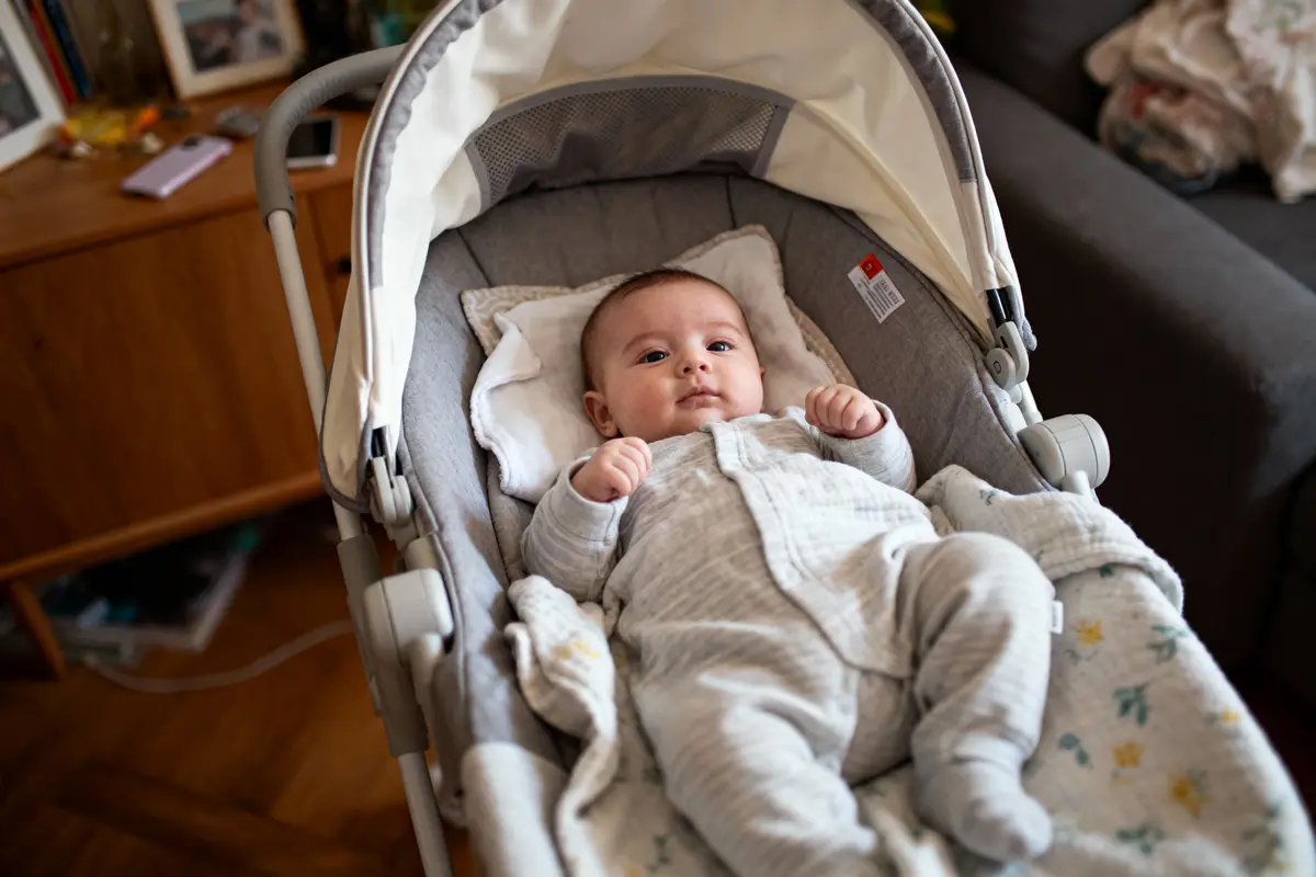 Bébé en pyjama gris allongé dans un landau, intérieur confortable avec meubles et objets divers autour.