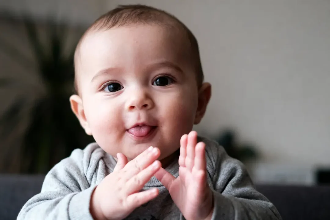 Bébé souriant avec un pull gris, les mains jointes, langue sortie, arrière-plan flou.