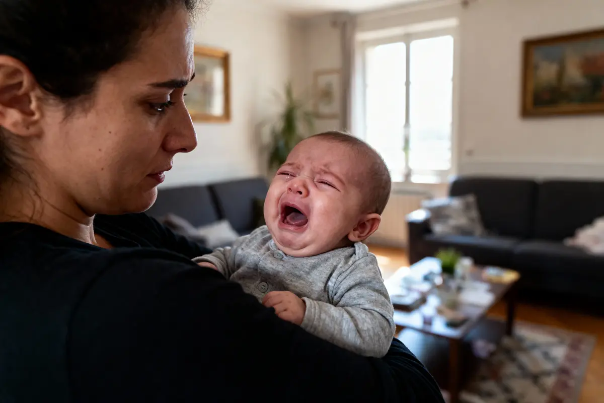 Femme tenant un bébé qui pleure, dans un salon lumineux et confortable.