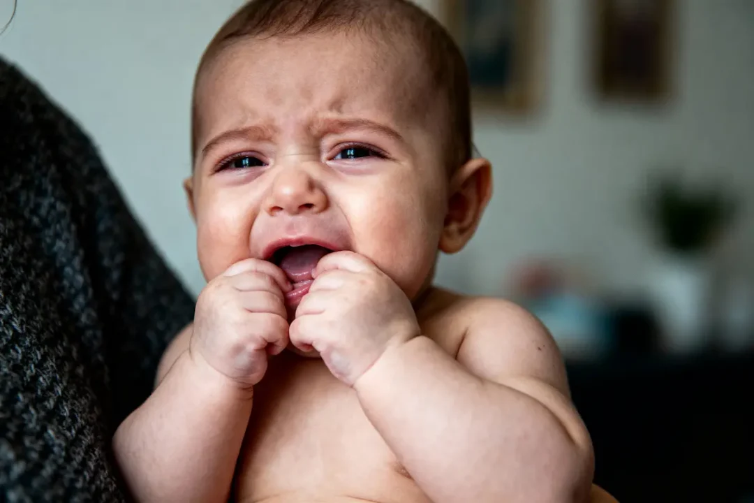 Bébé en pleurs avec les mains dans la bouche, fond flou et expression angoissée.