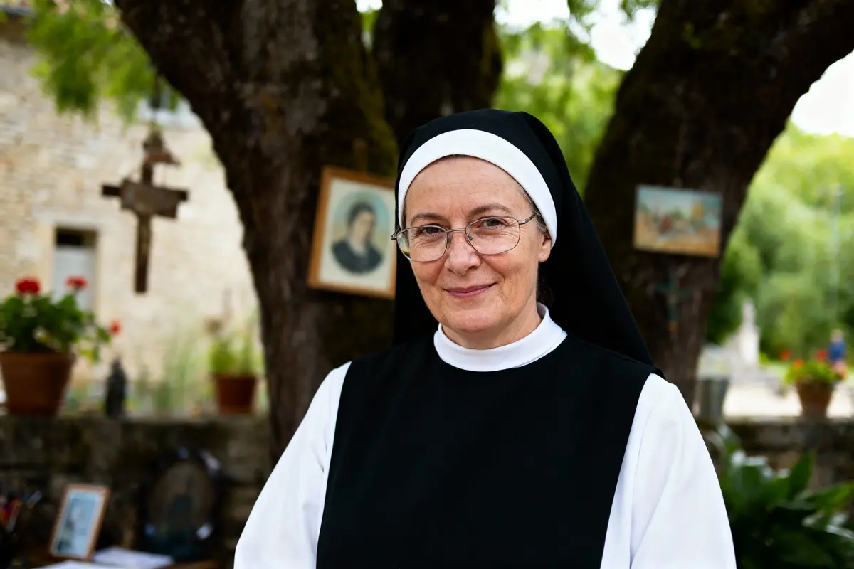 Religieuse souriante avec lunettes et voile noir, debout devant un arbre, décor extérieur religieux en arrière-plan.