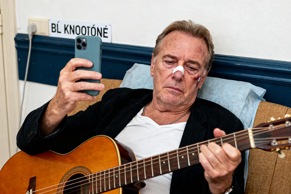 Homme blessé au visage, tenant un smartphone et jouant de la guitare dans un intérieur simple.