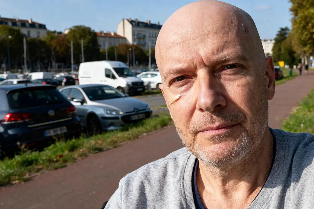 Un homme avec un pansement sur la joue marche près d'une route avec des voitures garées.