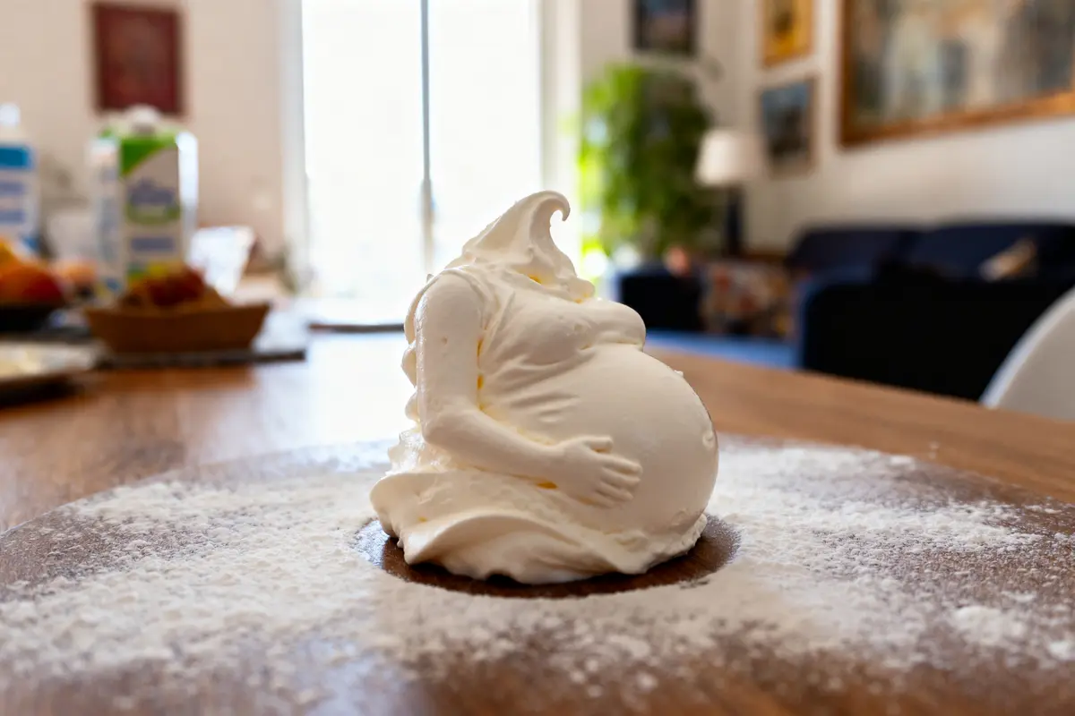 Figurine de crème fouettée en forme de femme enceinte, posée sur une table en bois, intérieur lumineux.