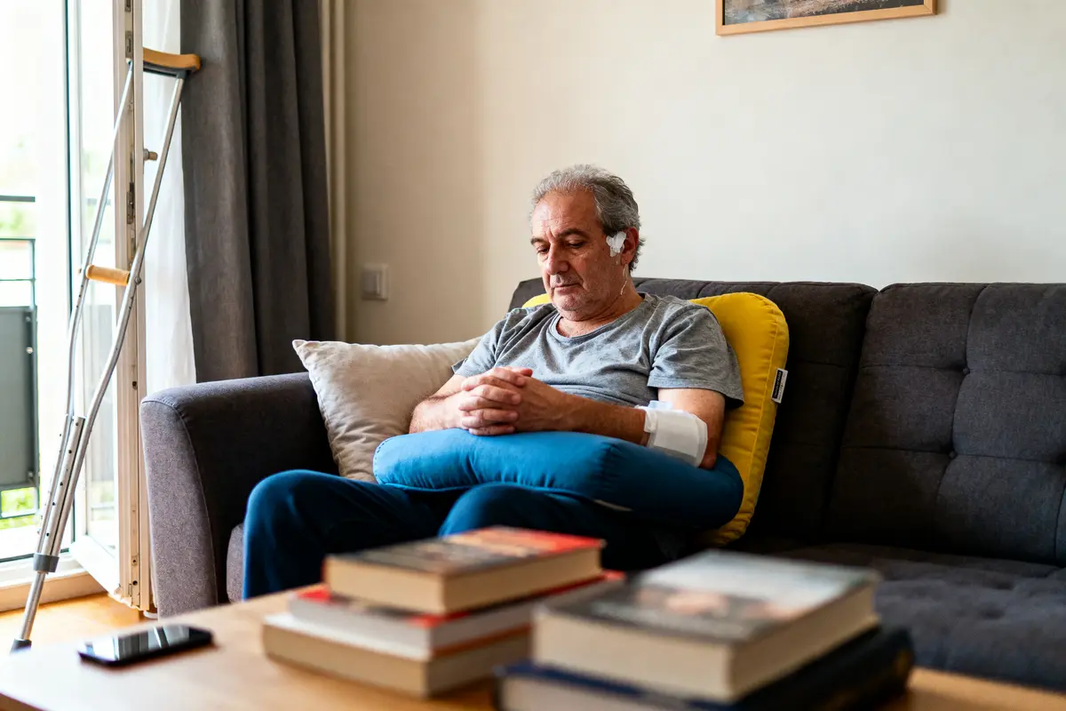 Homme blessé assis sur canapé, appuyé sur coussin. Béquilles, livres et téléphone en arrière-plan.