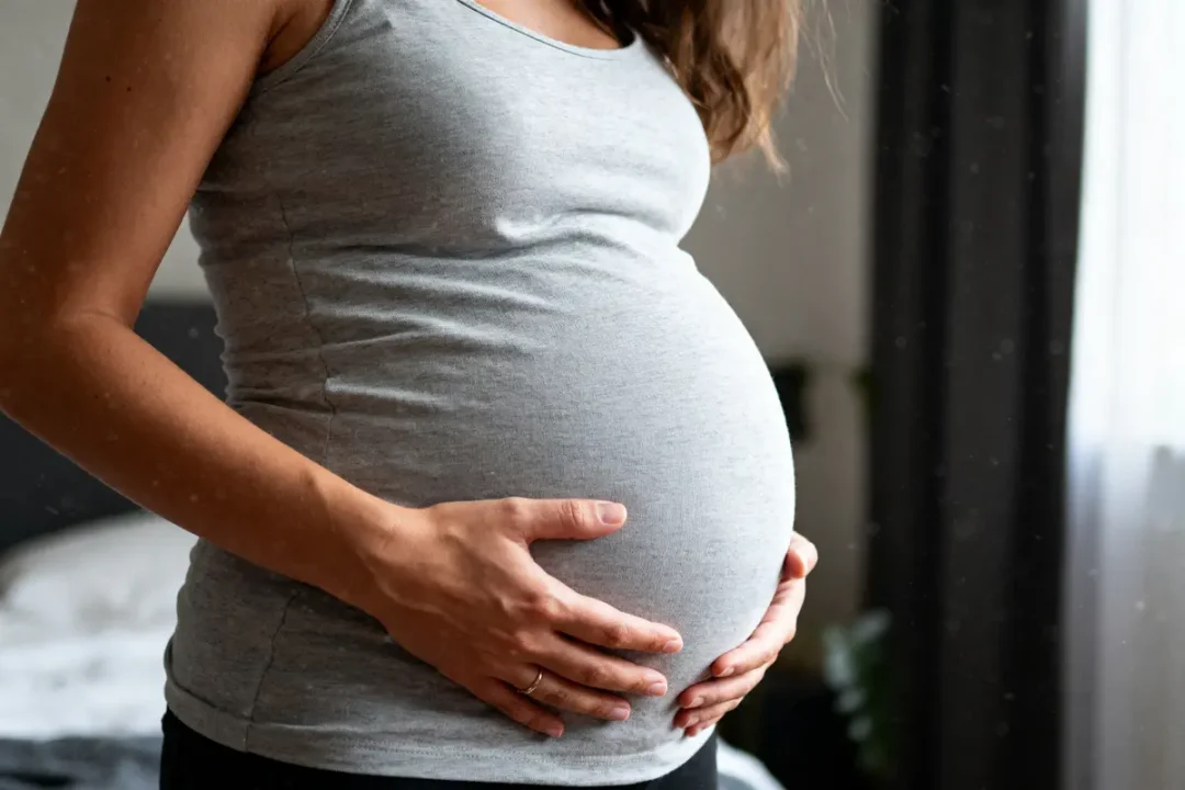 Femme enceinte touchant son ventre, portant un débardeur gris, intérieur avec lumière naturelle.