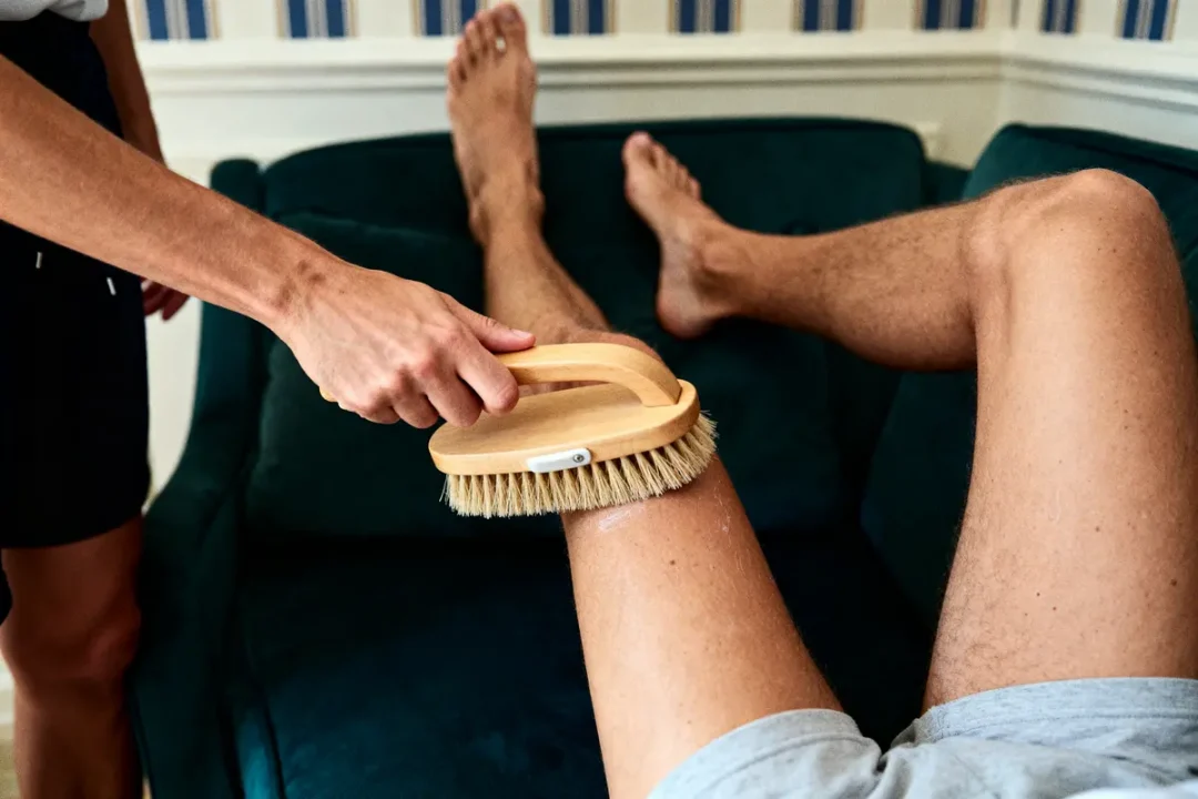 Personne brosse la jambe d'une autre avec une brosse en bois, sur un canapé vert.