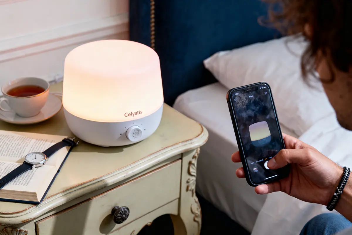 Lampe Celyatis sur table de chevet, smartphone, livre, montre et tasse de thé à proximité.