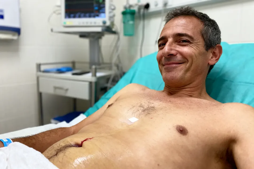 Homme souriant dans un lit d'hôpital, cicatrice et monitorage visible en arrière-plan.