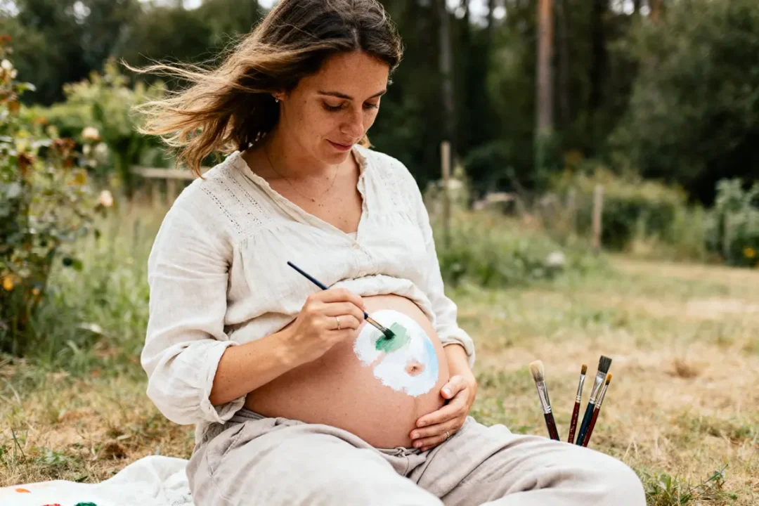 Femme enceinte peignant sur son ventre dans un jardin, entourée de végétation.