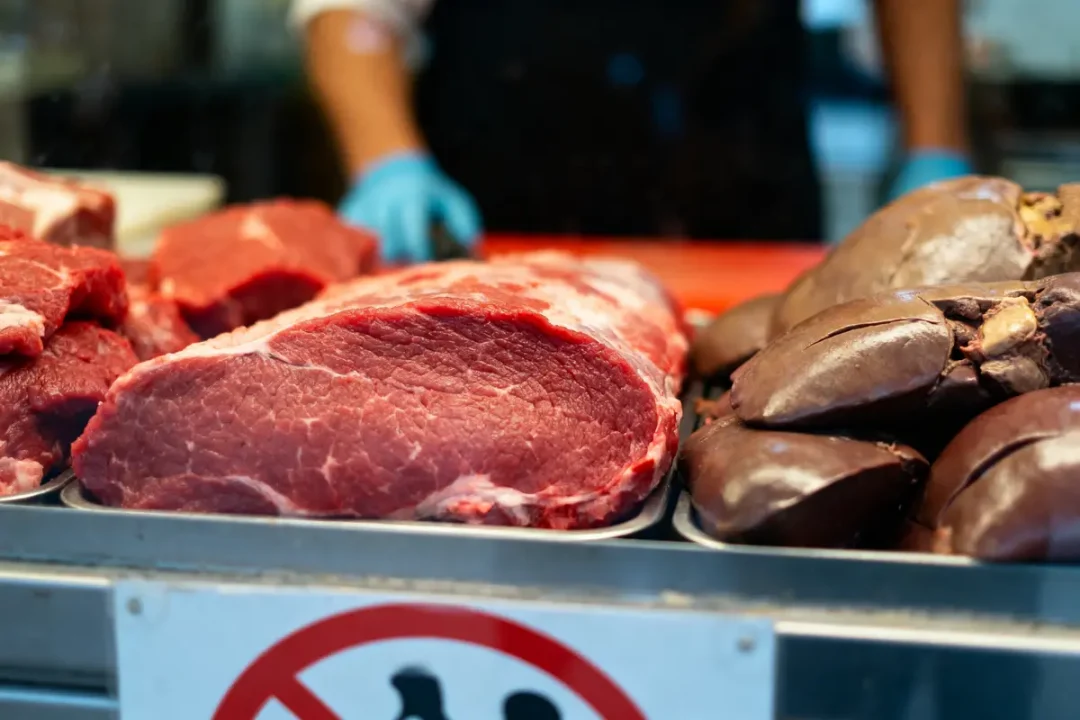 Morceaux de viande crue sur un étal de boucherie, avec une personne portant des gants bleus en arrière-plan.