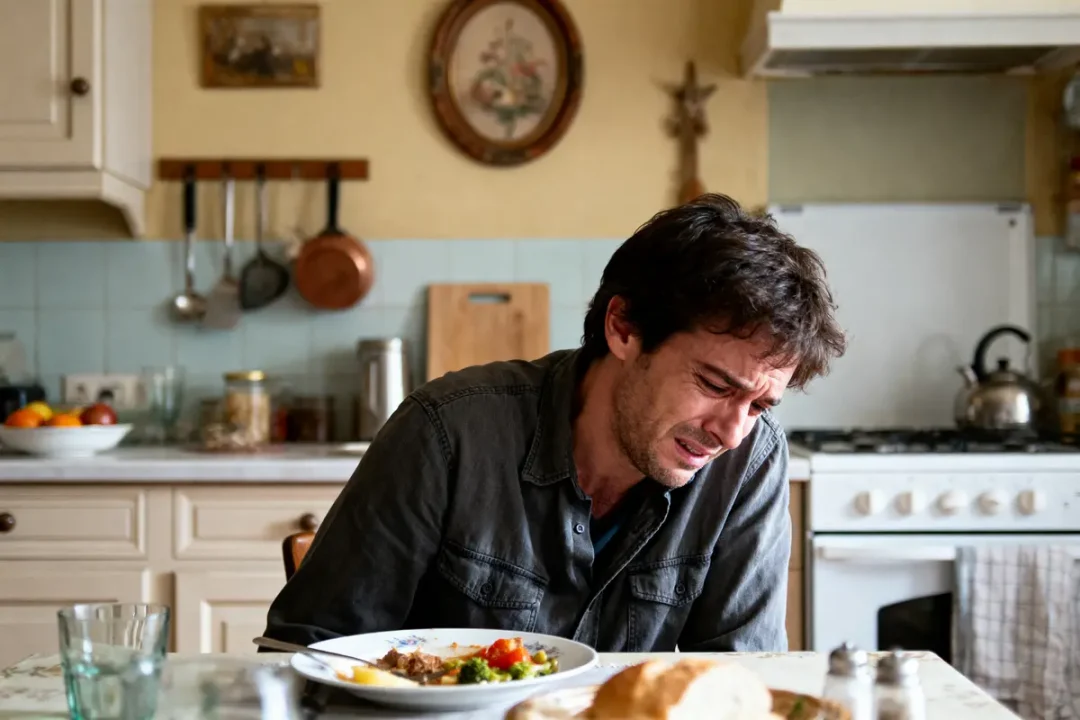 Un homme triste assis à une table dans une cuisine, assiette partiellement vide devant lui.