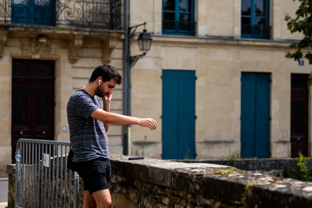 Un homme écouteurs, appuyé contre une rambarde, regarde son téléphone, devant un bâtiment en pierre.
