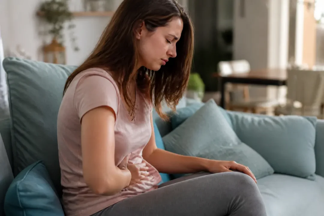 Femme assise sur un canapé, main sur le ventre, exprimant une douleur ou un inconfort abdominal.