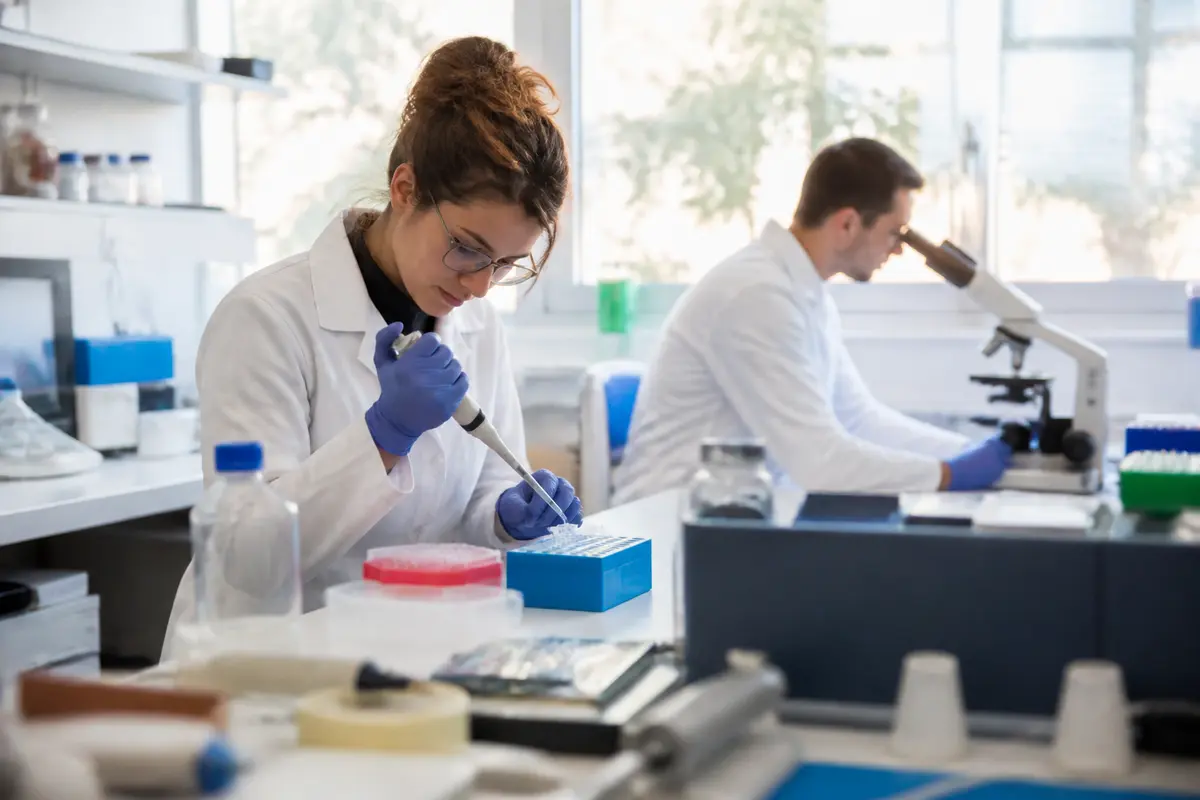 Deux scientifiques en laboratoire, une femme pipette, un homme au microscope, tous deux concentrés et en blouse blanche.