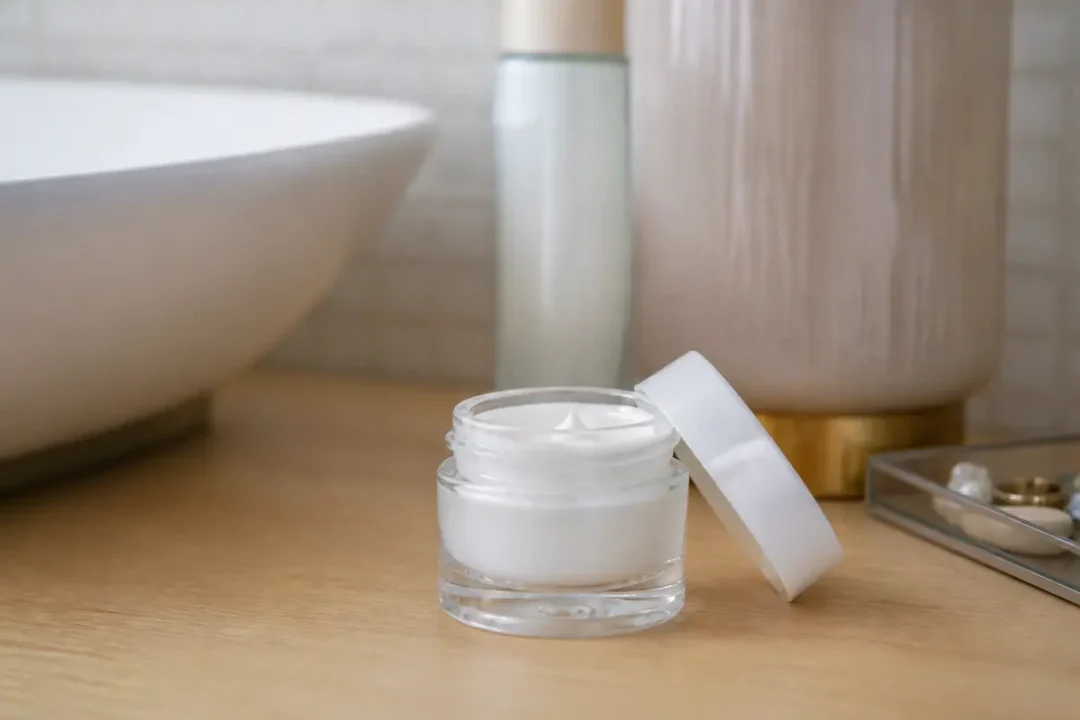 Pot de crème blanc ouvert sur une table en bois, avec accessoires de salle de bain.