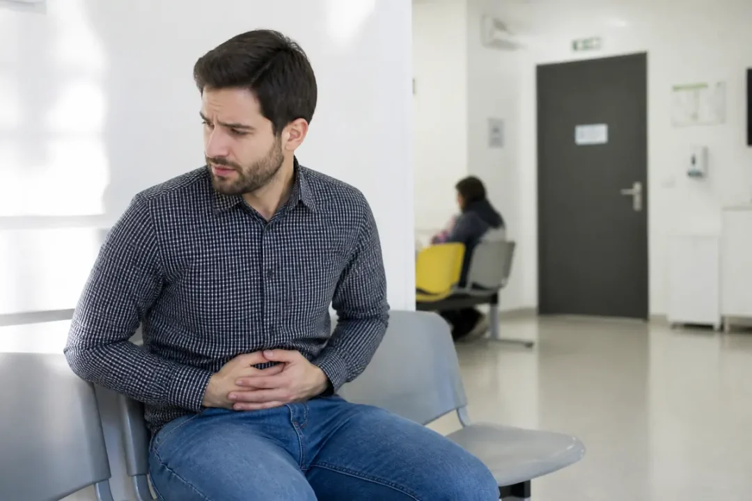 Un homme en salle d'attente, mains sur le ventre, souffrant, assis sur une chaise grise.