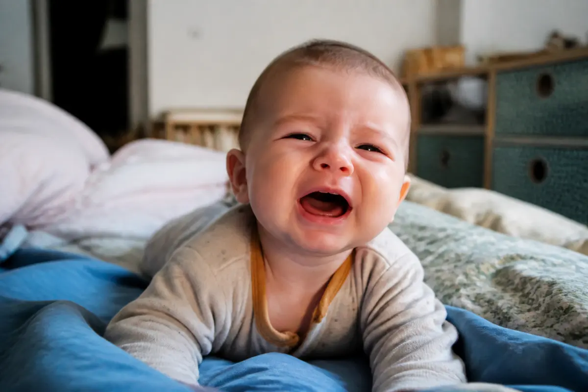 Un bébé allongé sur un lit, pleurant, vêtu d’un pyjama gris clair.
