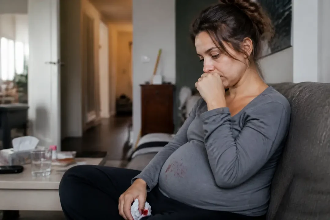 Femme enceinte assise, pensive, regardant vers le bas, sur un canapé dans un salon moderne.