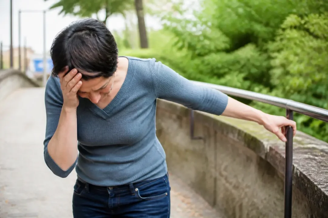 Une femme tenant une rampe en se tenant la tête, semble éprouver un malaise ou des étourdissements dehors.